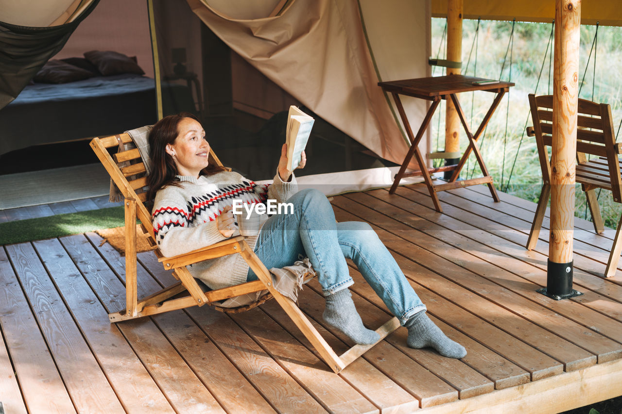 Brunette woman in nordic sweater reading book and relaxing in glamping in nature