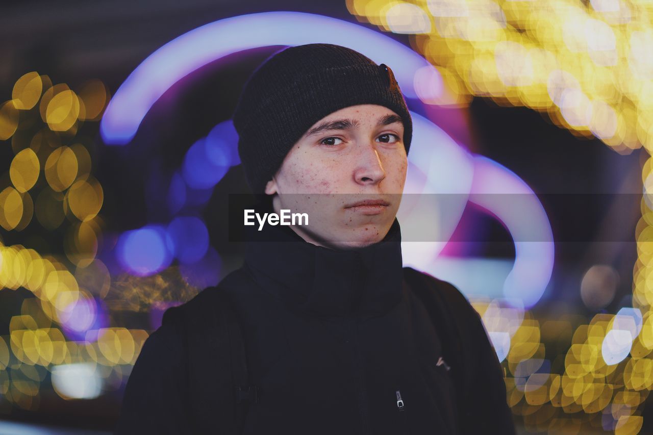 PORTRAIT OF YOUNG MAN STANDING AGAINST ILLUMINATED LIGHT