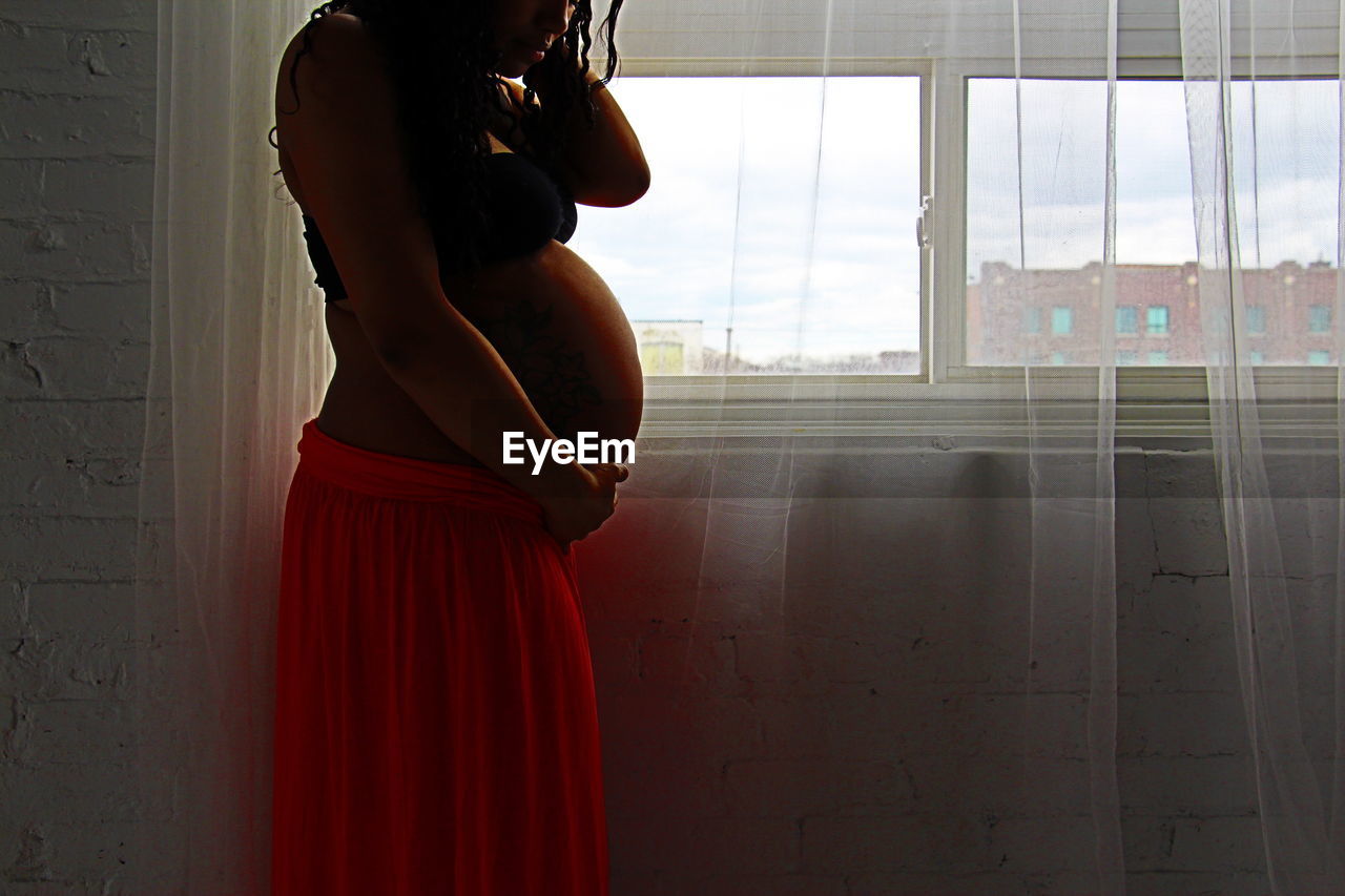 Midsection of pregnant woman standing by window at home