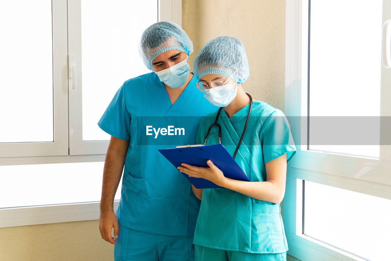 Serious male and female medics in masks reading report on clipboard while standing in corridor in clinic