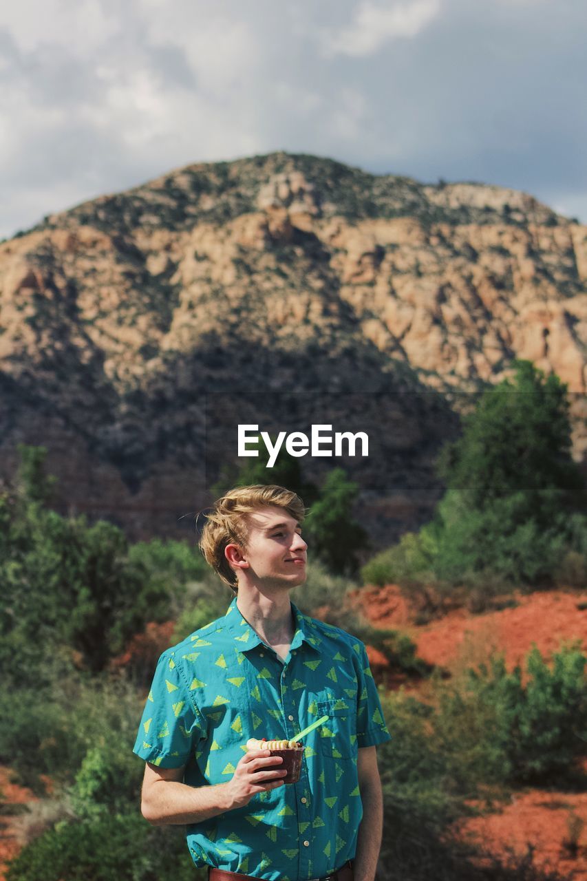Young man holding sweet food looking away while standing against mountain