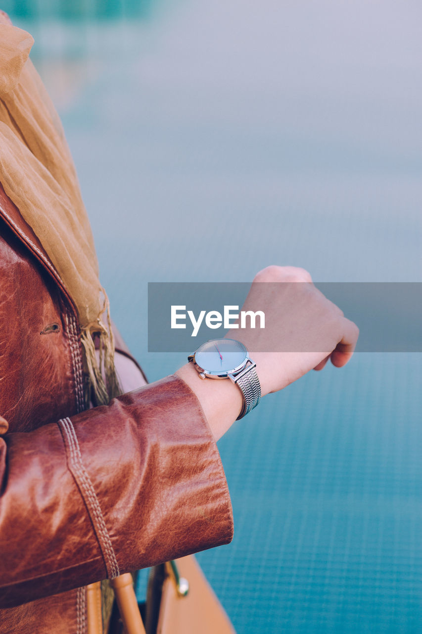 Midsection of woman checking time on wristwatch standing against blue wall