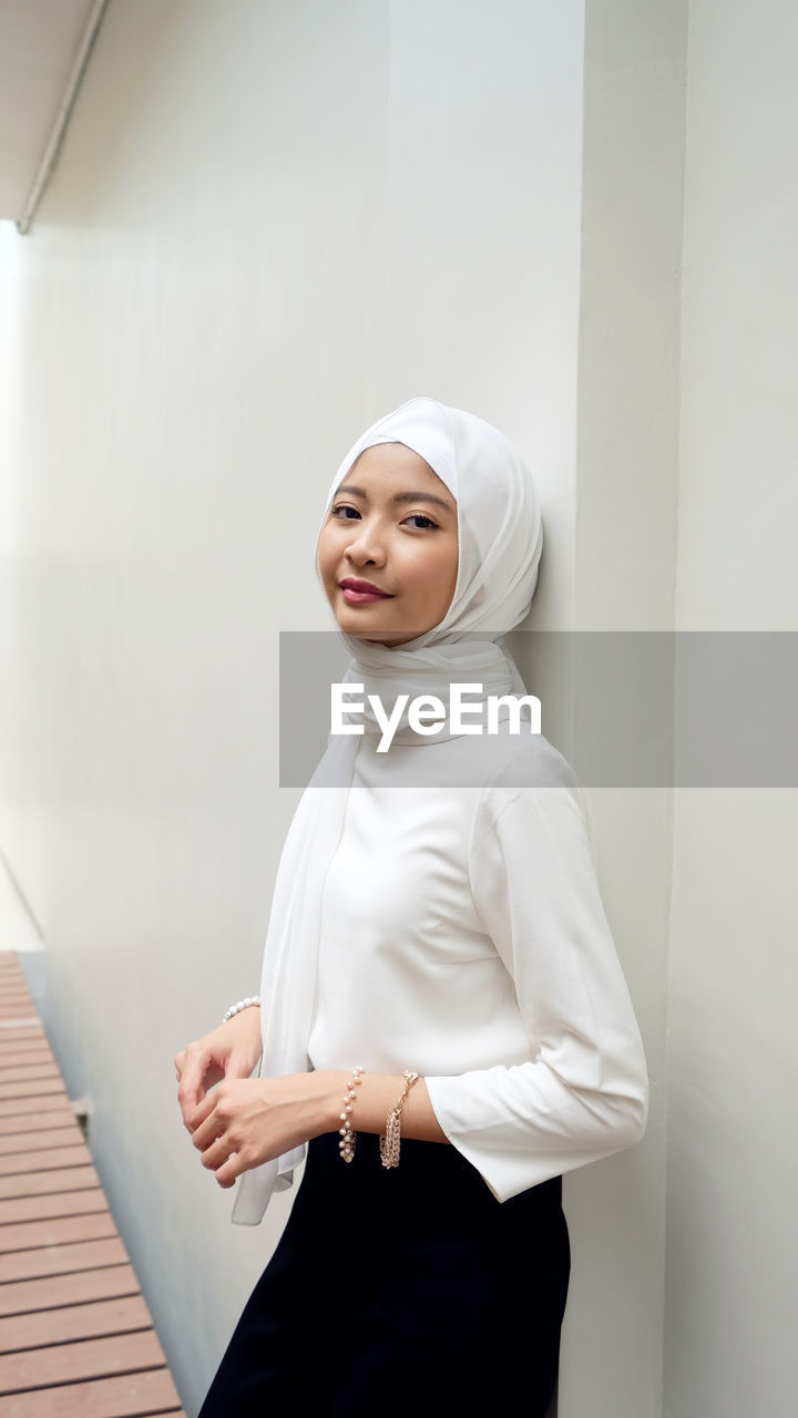 SMILING YOUNG WOMAN STANDING AGAINST WALL