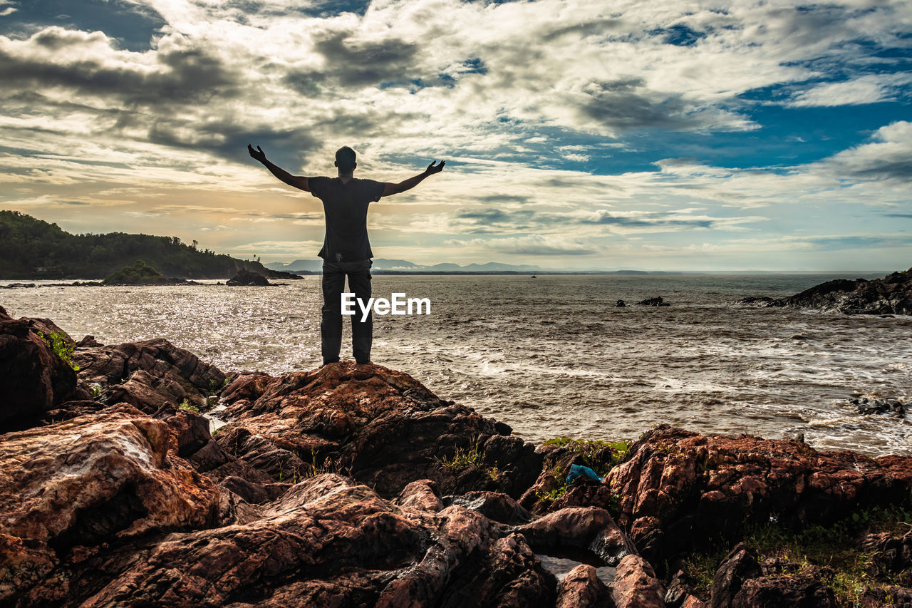 MAN STANDING ON ROCK AT SHORE AGAINST SKY