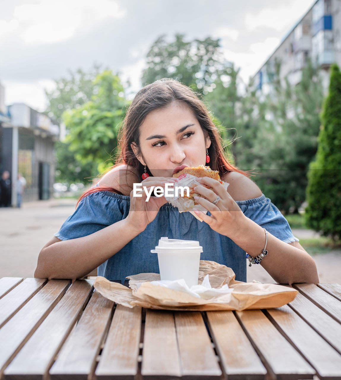 Hungry stylish woman, enjoying eating a burger outdoors, dressed in jeans shirt, wearing sunglasses