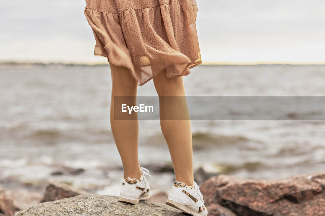 A young woman stands on the rocky shore of the sea. a dress flying in the wind close-up.