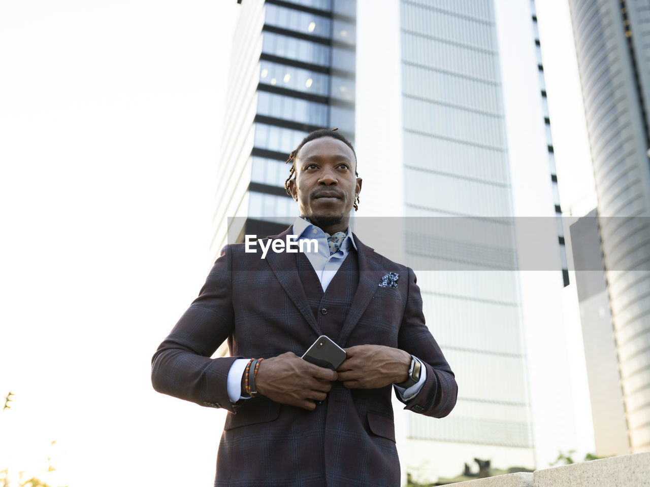 Low angle of serious african american male entrepreneur wearing classy suit standing with smartphone in hand on street in city center and looking away
