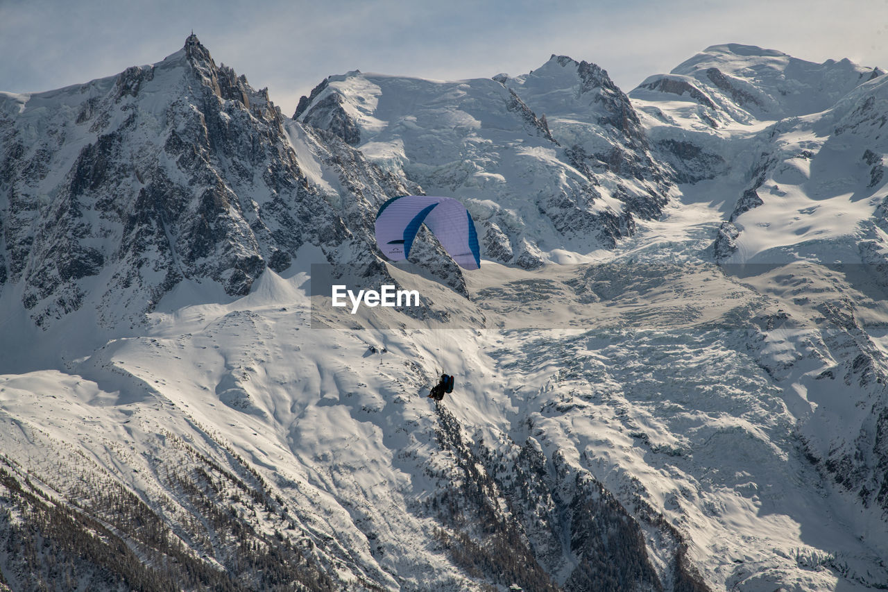Person paragliding over snow covered mountains against sky