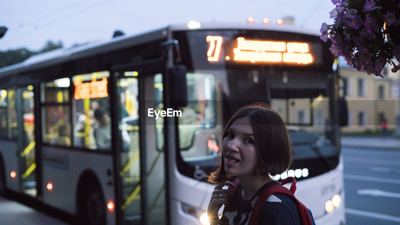 Portrait of young woman sticking out tongue against bus in city