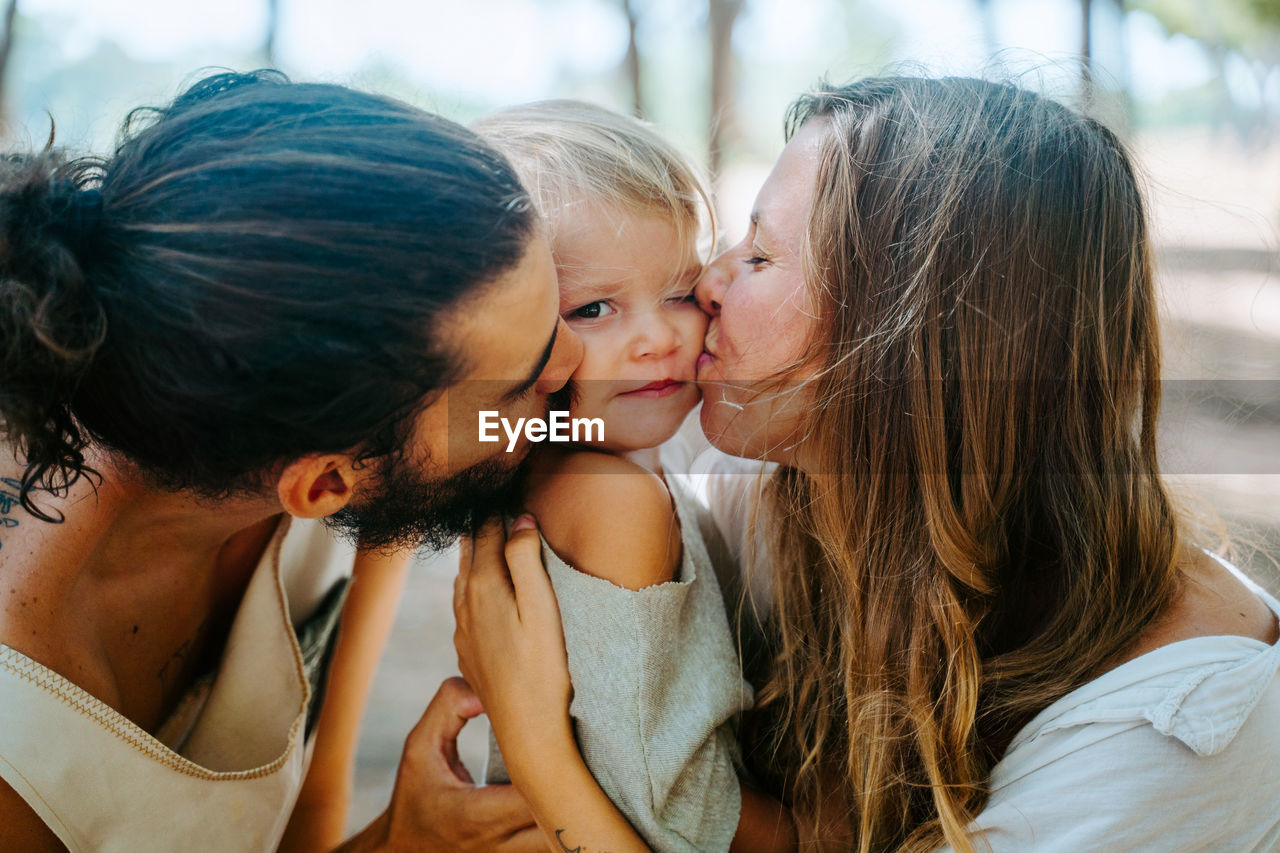 Side view of multiethnic mother and father cuddling and tenderly kissing little child in cheek while resting together in forest