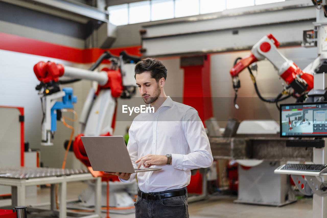Engineer using laptop standing in robot factory