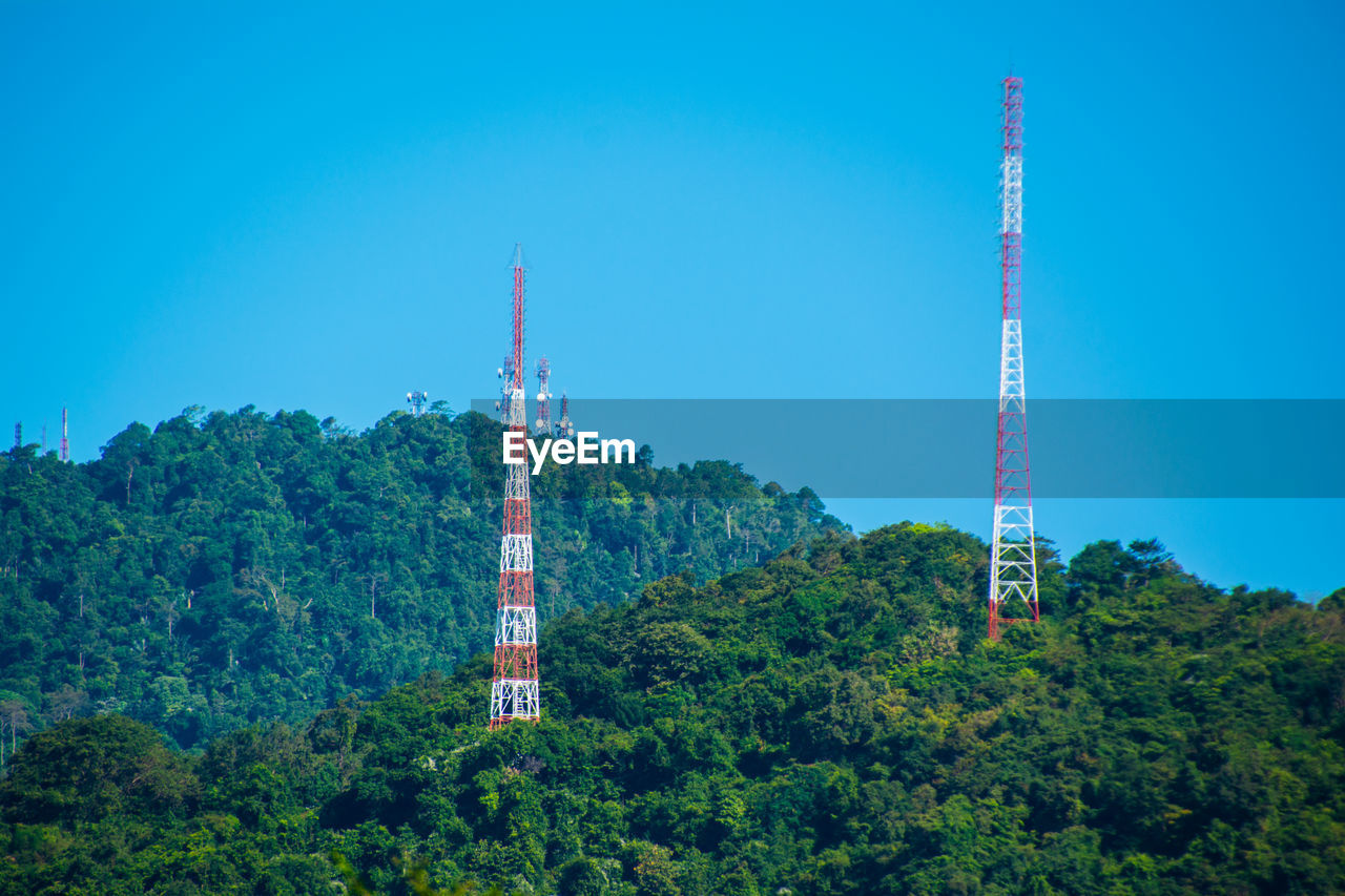 Low angle view of communications tower and trees against clear blue sky