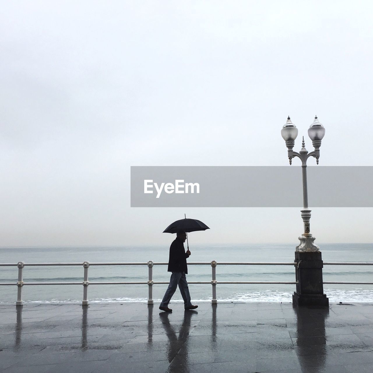 Side view of man walking under umbrella during rainfall by sea against sky