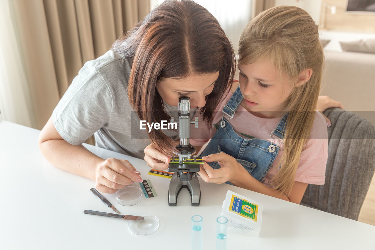 Woman looking through microscope sitting by daughter at home