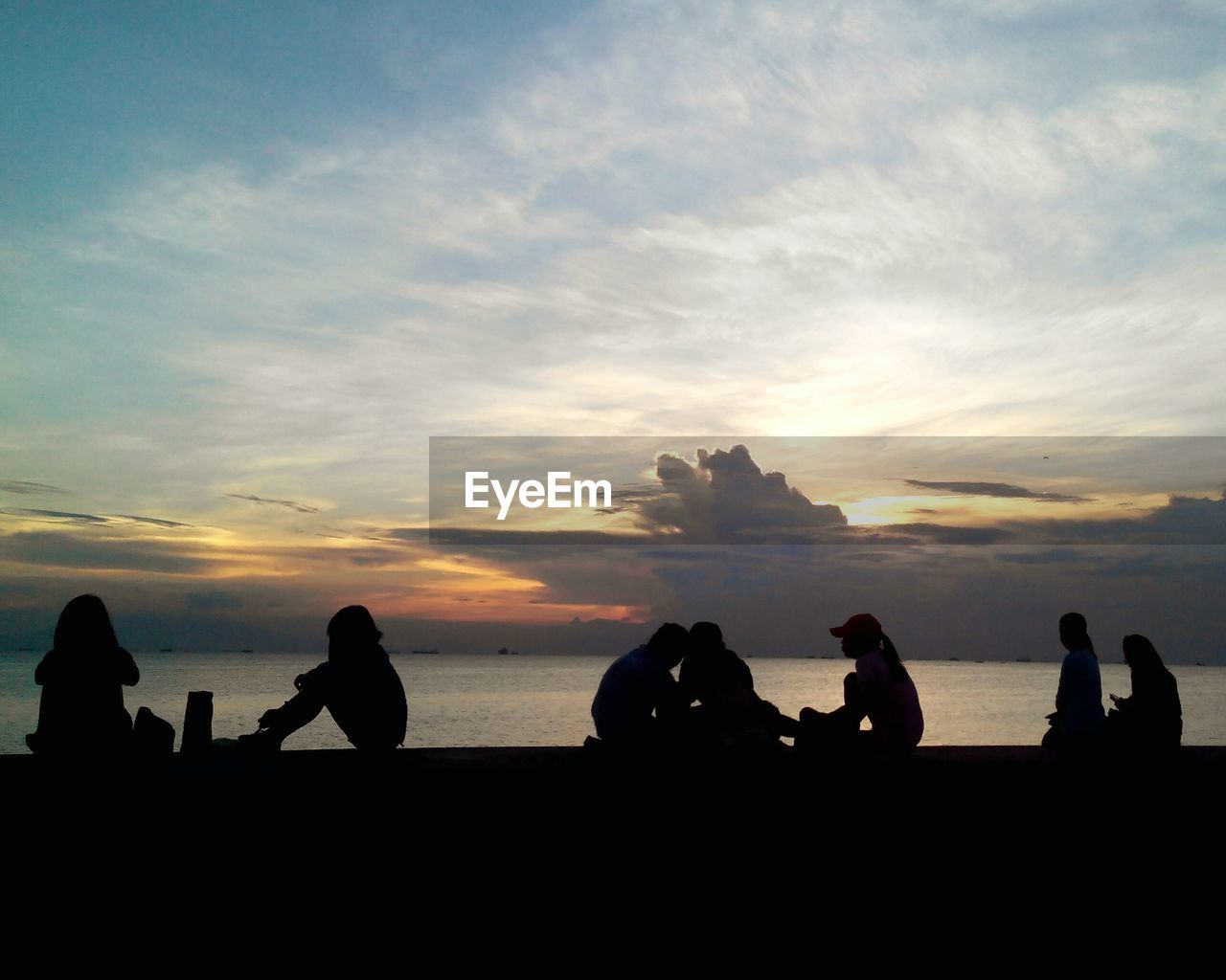 Silhouette people sitting on retaining wall by sea against cloudy sky