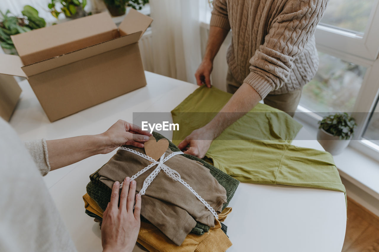 Fashion designer with colleague putting heart shaped card on folded clothes in workshop