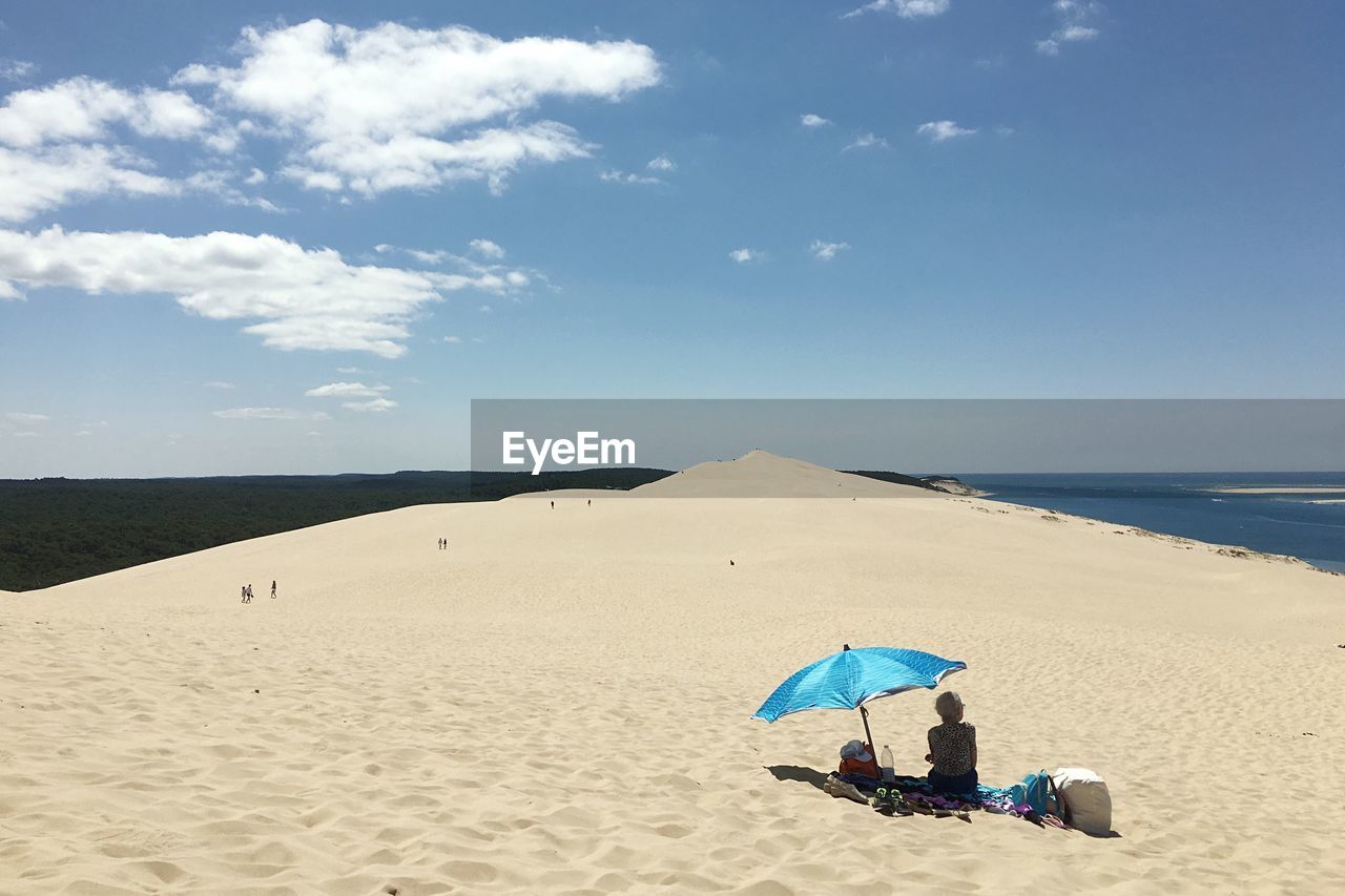 Rear view of woman sitting by umbrella at beach against sky