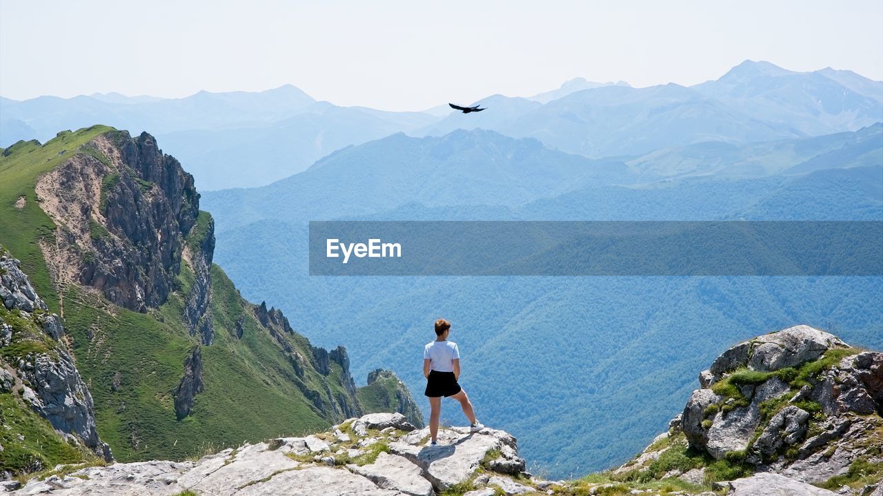 A woman looks at the landscape from the top of a mountain and a bird flies over her head