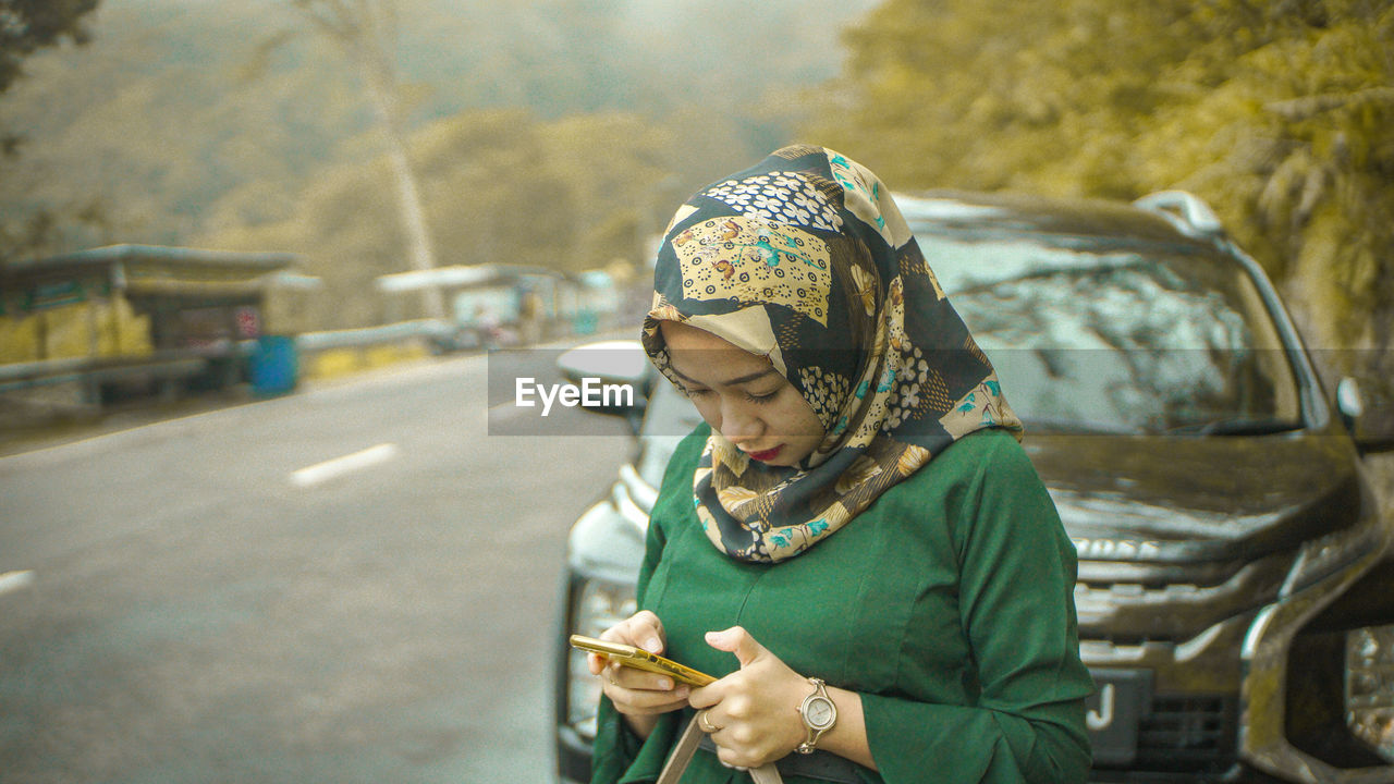 Young woman in hijab using mobile phone by car on road
