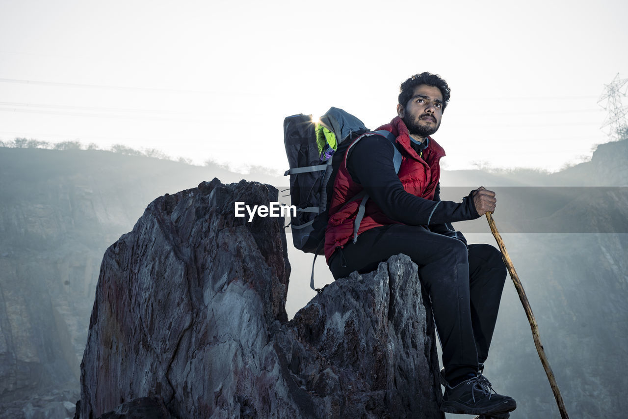 Young indian mountaineer sitting on the top of the mountain on a cliff with a backpack and a stick.