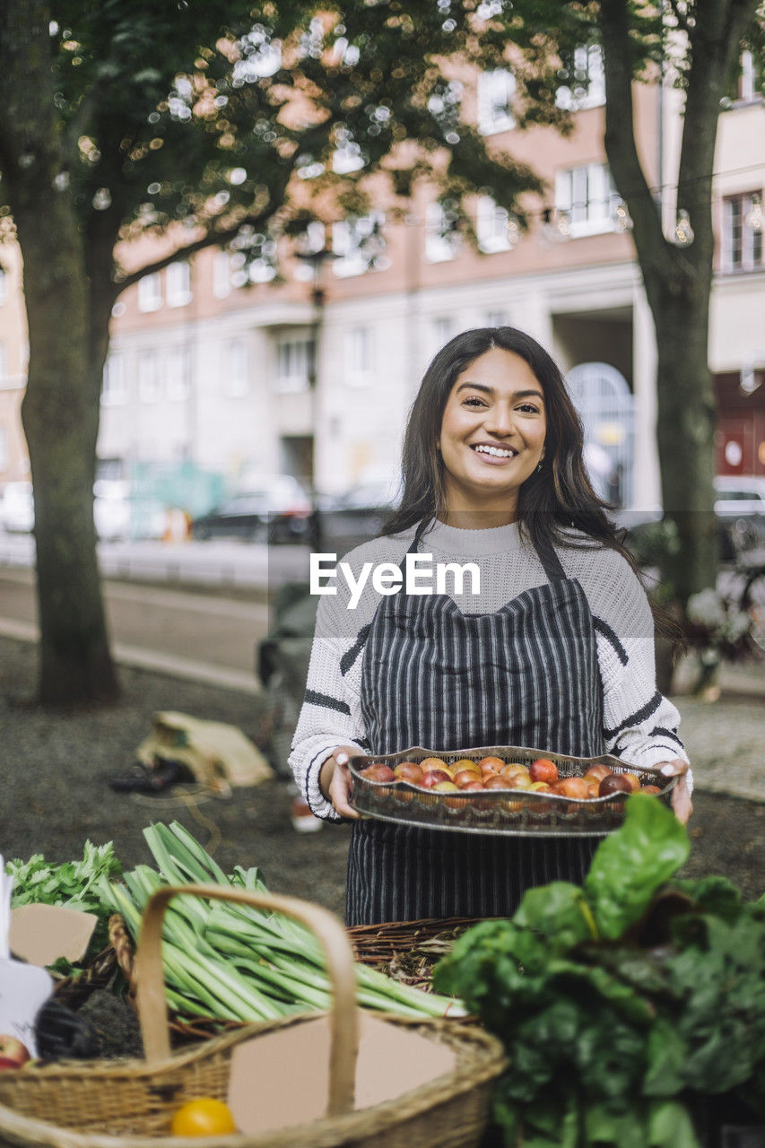 Portrait of young female vendor holding vegetable tray near stall at farmer's market