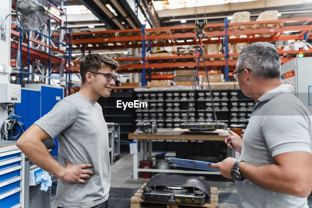 Smiling male coworker looking at mature engineer in illuminated factory