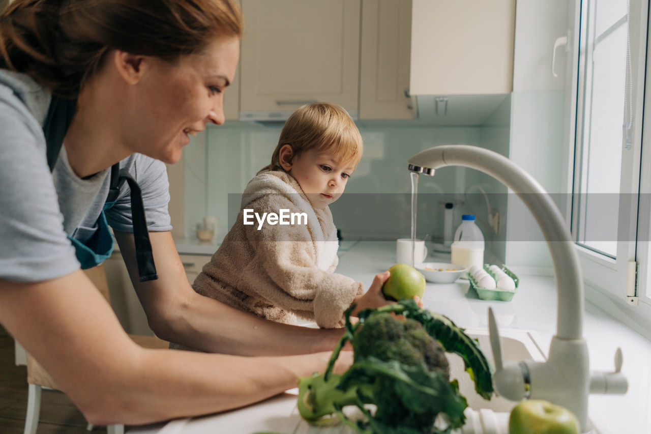 Mother and little child toddler are washing vegetables together in the kitchen for cooking dinner.