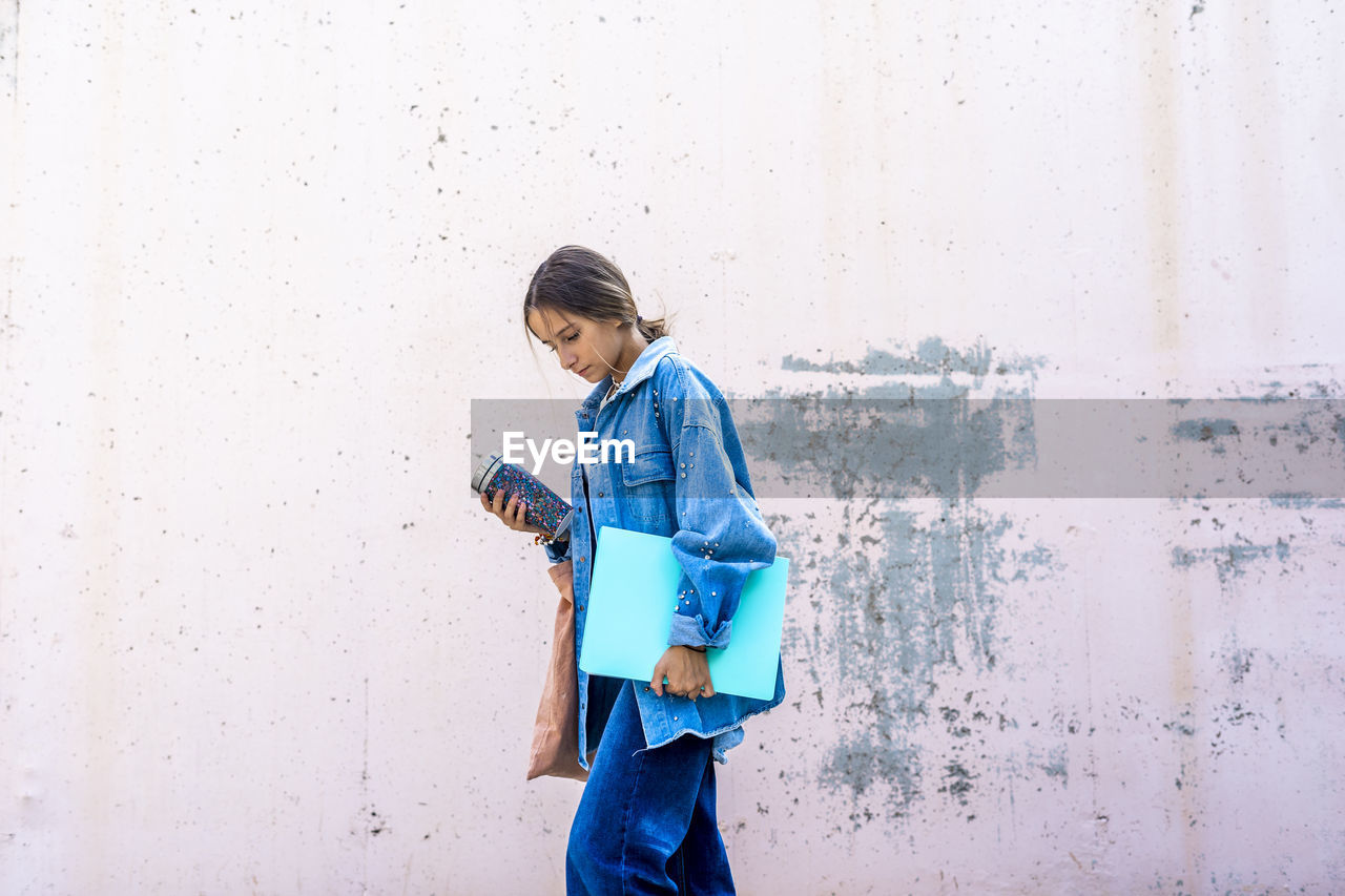 Teenage girl walking with book and bottle by wall on sunny day