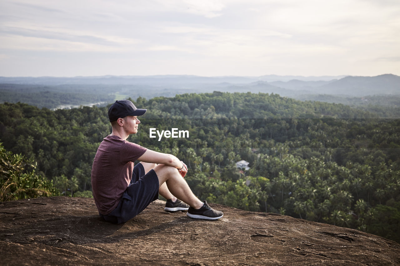 Pensive man resting at the edge of a rock and looking at view. landscape with tropical rainforest.