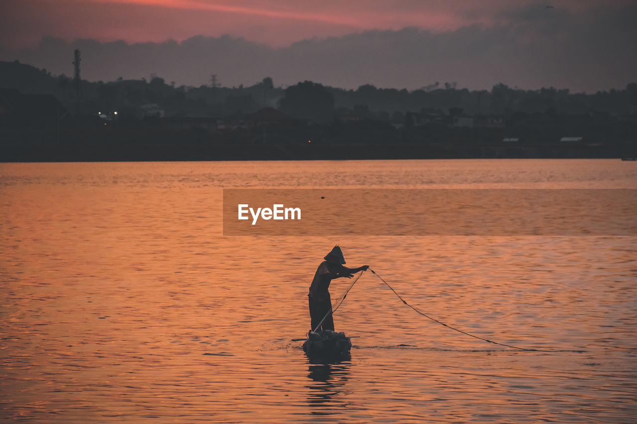 Silhouette man fishing in sea against orange sky