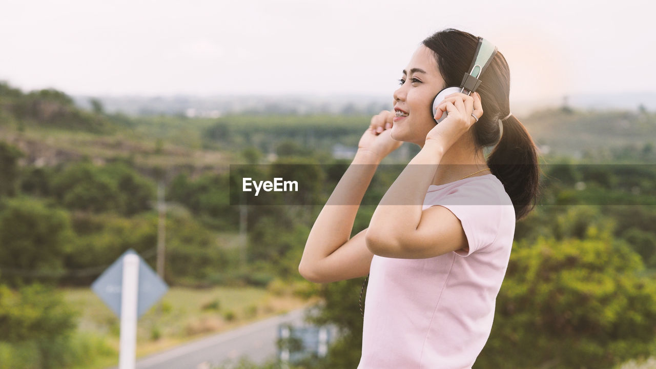 Side view of smiling young woman listening music against sky