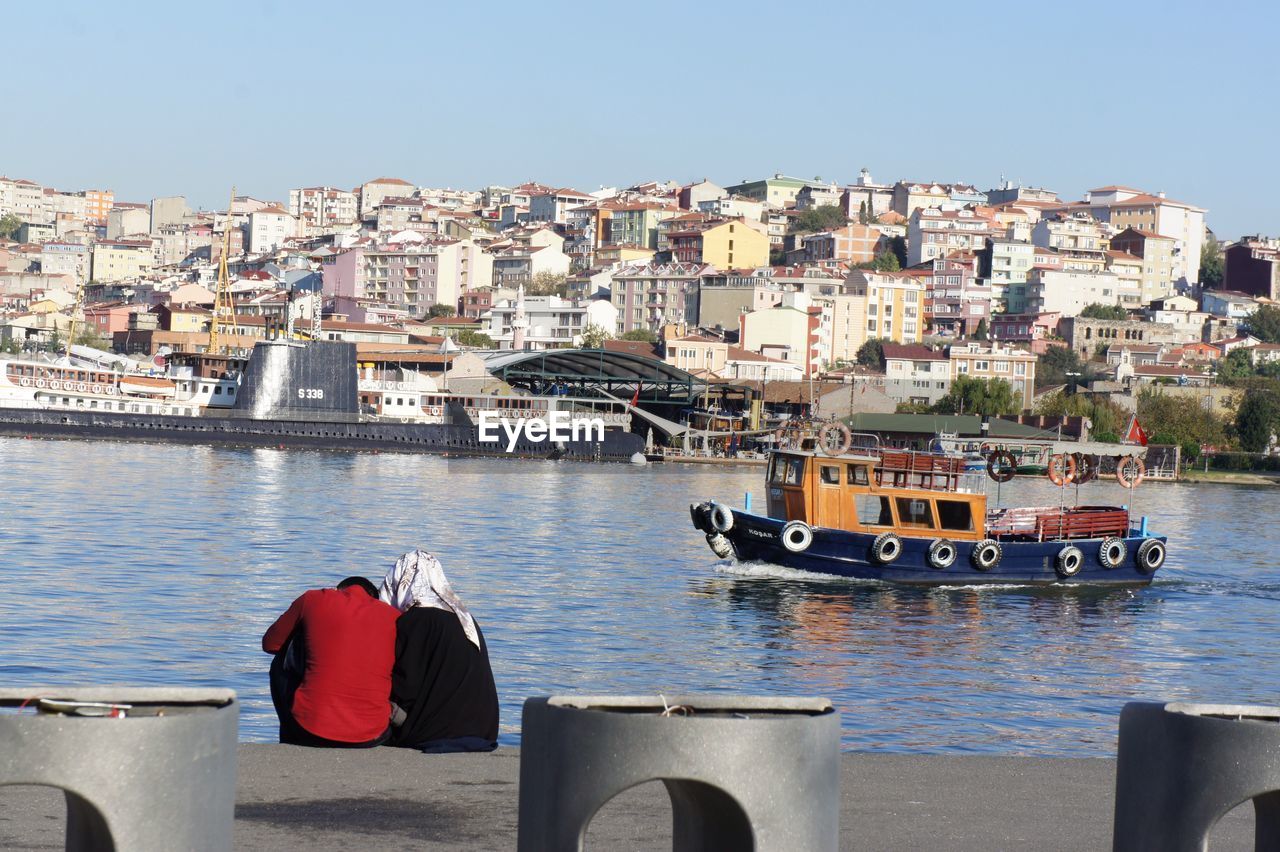 Rear view of couple sitting by canal against boat and buildings on sunny day in city