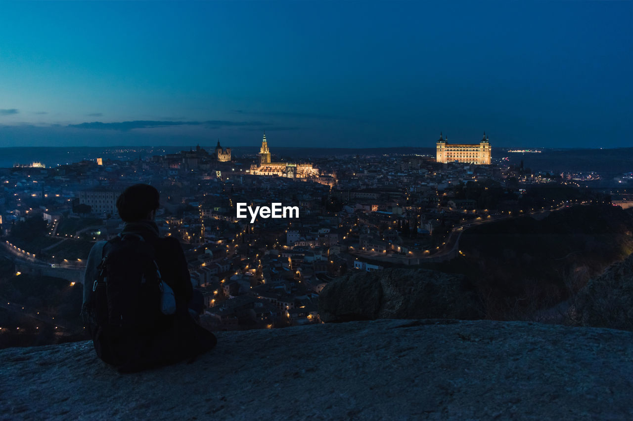 Back view of silhouette of male traveler sitting on hill and enjoying spectacular night cityscape of old city toledo in spain with illuminated medieval buildings