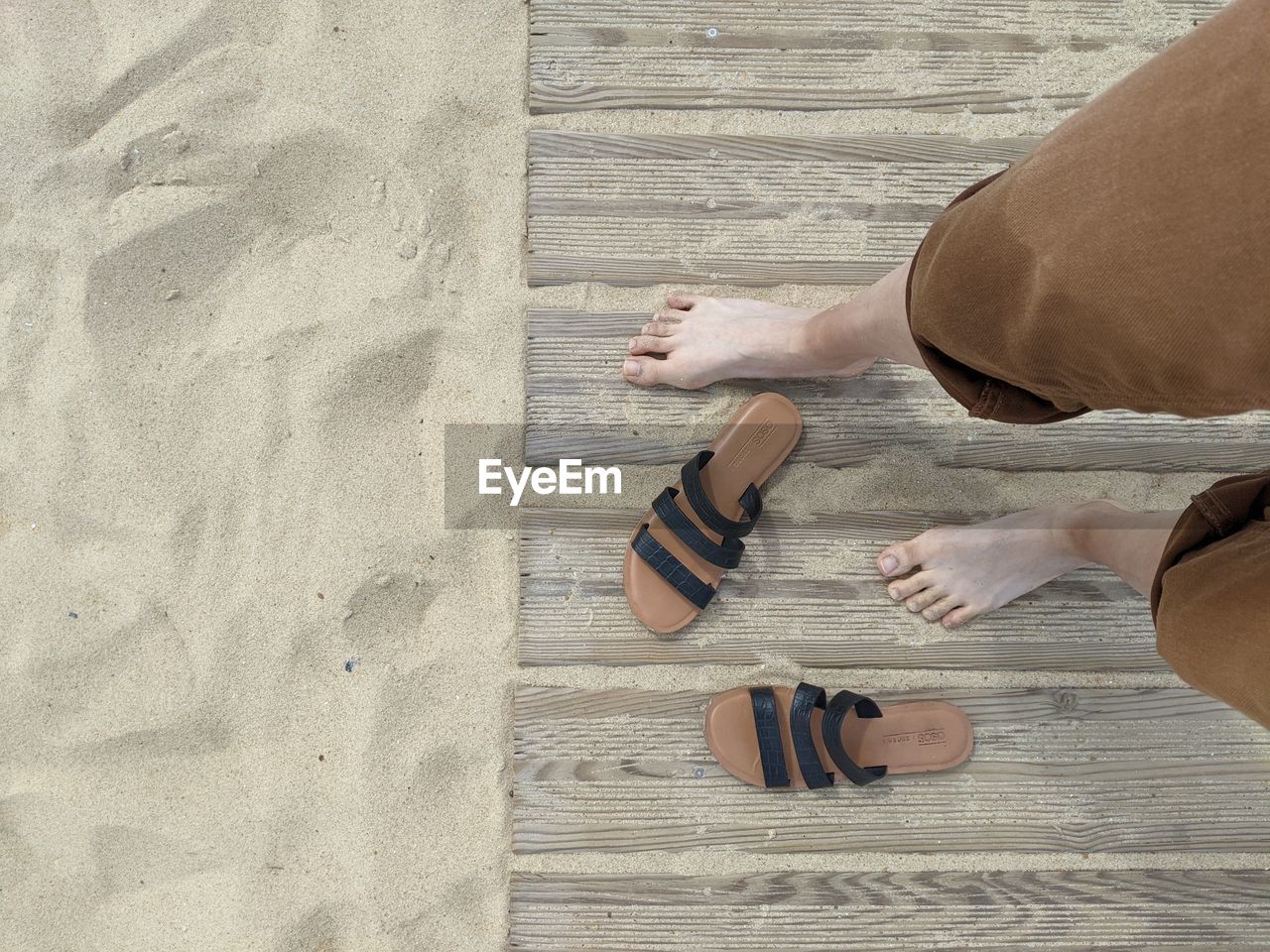 Personal perspective of looking down at bare feet with sandals on sand.