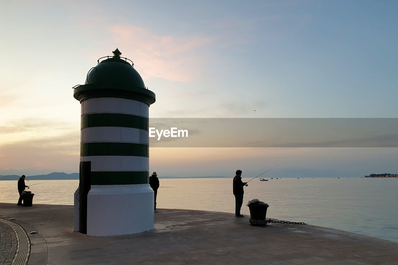 lighthouse, sea, sky, water, tower, beach, ocean, architecture, guidance, sunset, coast, built structure, nature, horizon, land, security, protection, building exterior, silhouette, cloud, horizon over water, travel destinations, building, scenics - nature, beauty in nature, travel, tranquility, men, coastline, shore, outdoors, trip, dusk, sunlight, vacation, tranquil scene, standing, pier, full length, bay, adult, tourism, holiday, leisure activity, lifestyles, idyllic