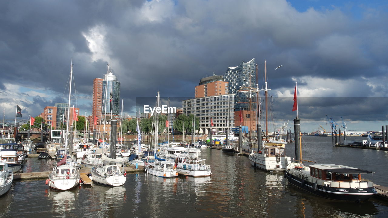 Boats moored at harbor by buildings against cloudy sky
