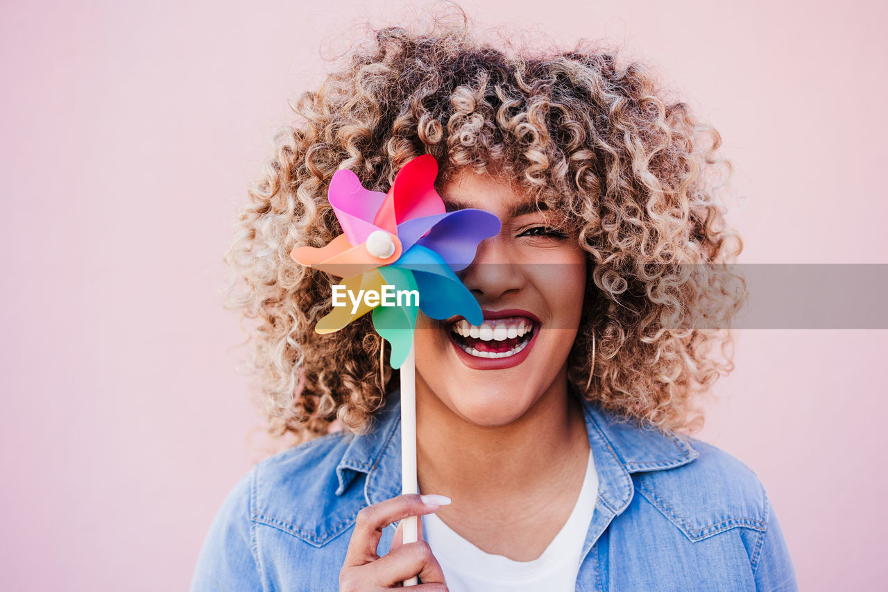 Beautiful happy hispanic woman with afro hair holding colorful pinwheel. pink background,wind energy