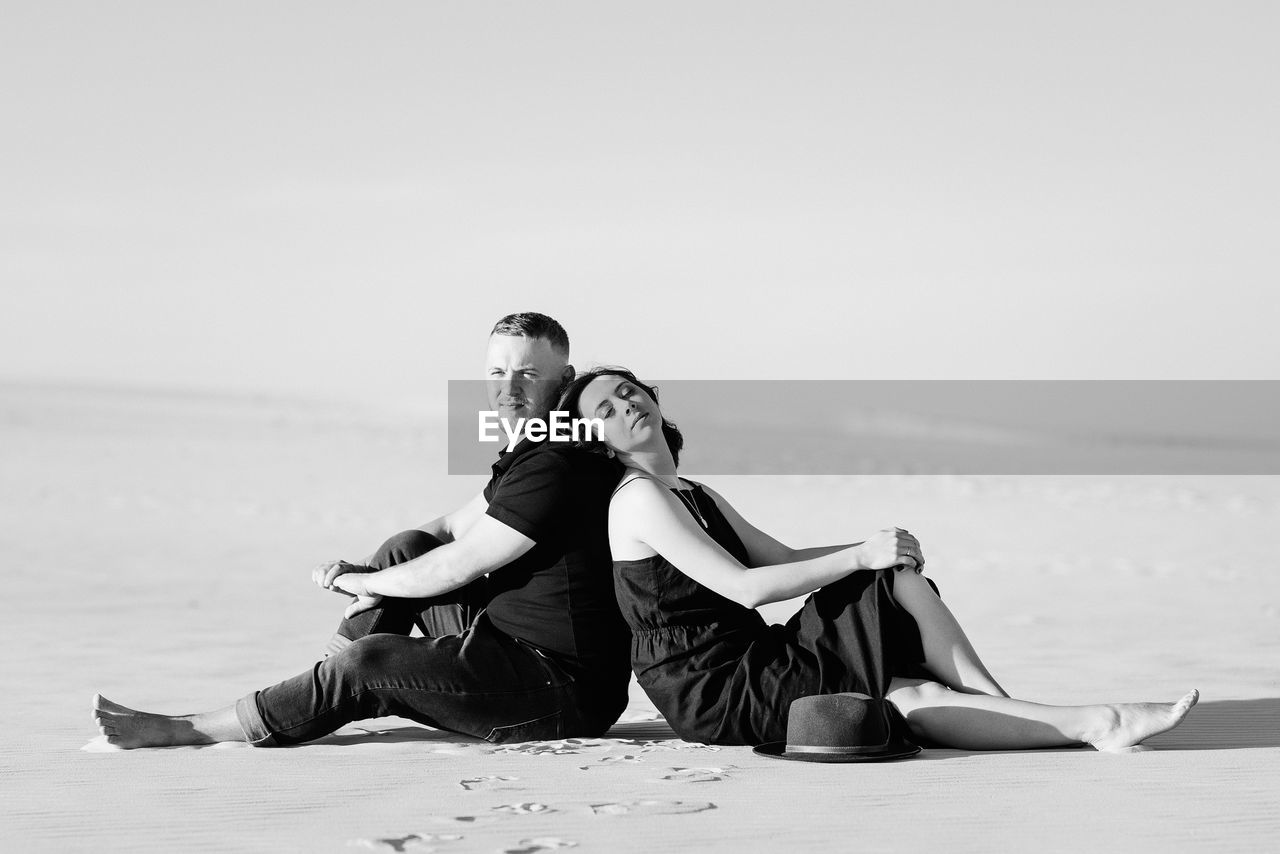 YOUNG COUPLE SITTING ON BEACH AGAINST SEA