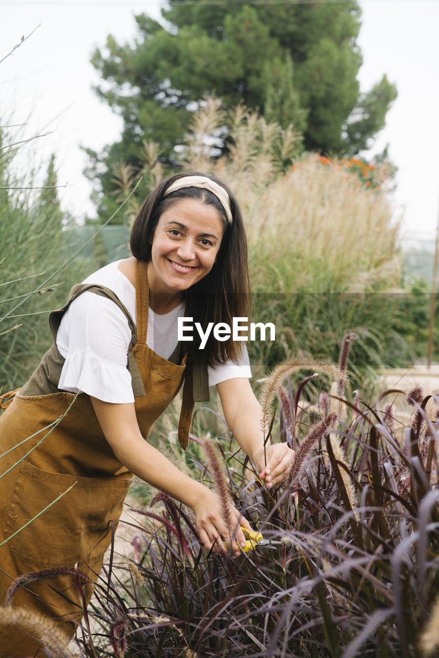Side view of positive adult ethnic female gardener with long dark hair in casual clothes and apron smiling while cutting reed plant in countryside