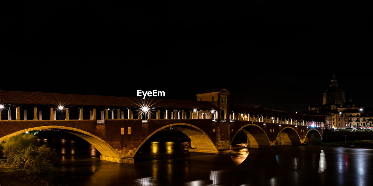 Illuminated bridge over river by buildings against sky at night in pavia, italy