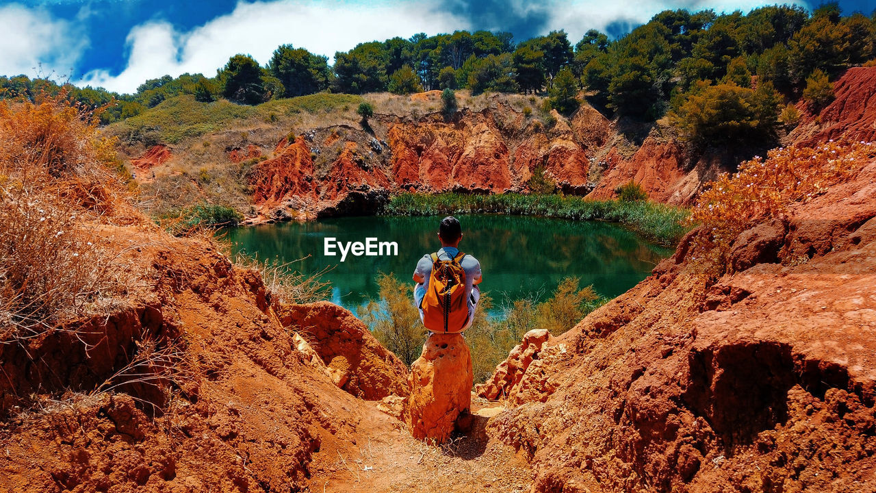 MAN STANDING ON ROCK BY LAKE AGAINST TREES