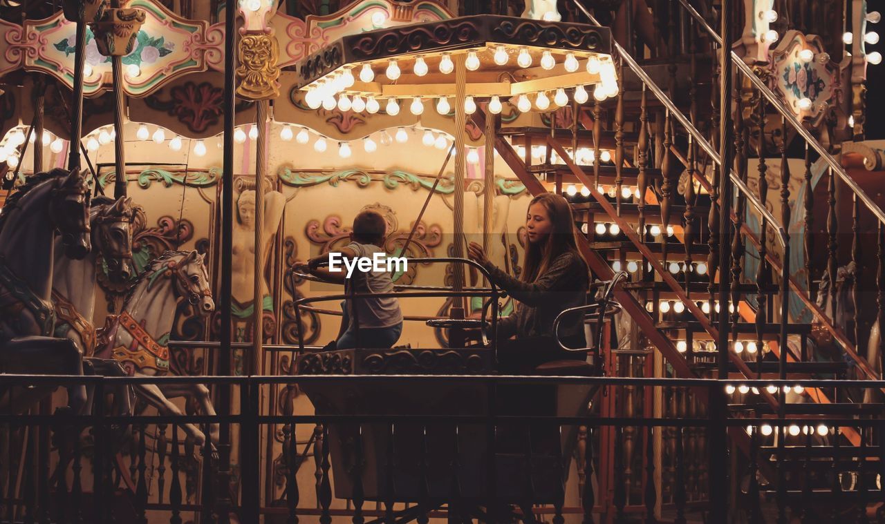 Siblings sitting in illuminated carousel horses at night