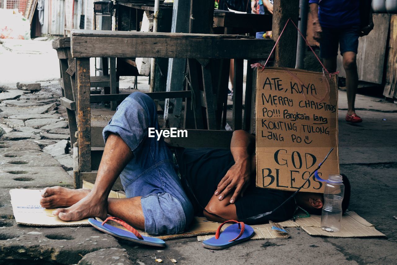 Homeless man lying on sidewalk by board with text