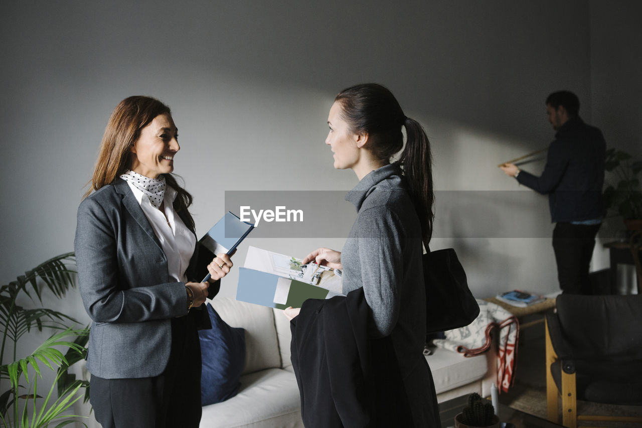 Happy woman and female realtor discussing brochure while man measuring wall in background