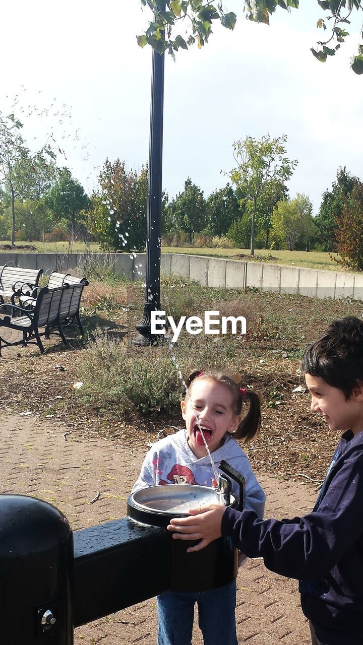 Siblings standing by drinking fountain spraying water