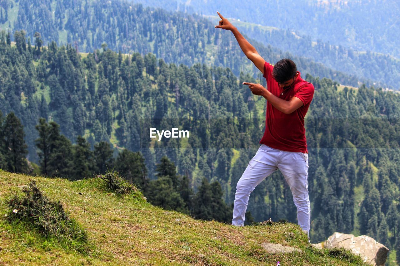 Full length of young man gesturing while standing on mountain against trees