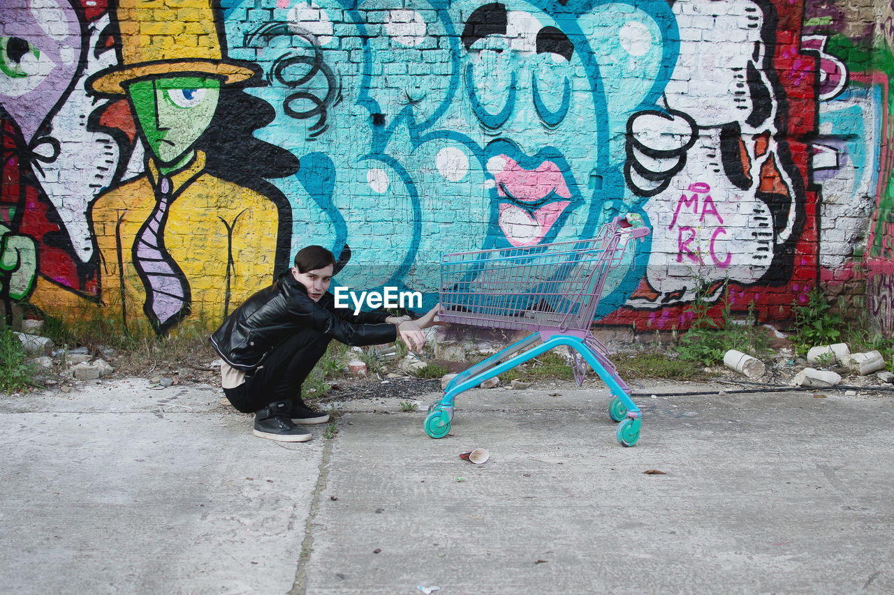 Side view of young man crouching by shopping cart against graffiti wall