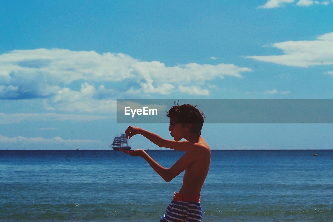 Shirtless boy standing at beach against sky