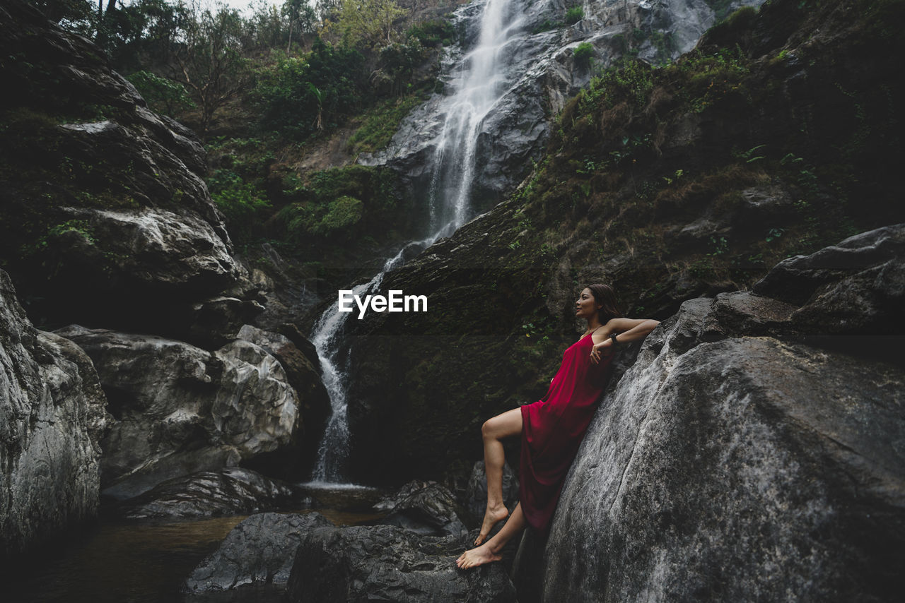 Side view of woman leaning on rock by waterfall