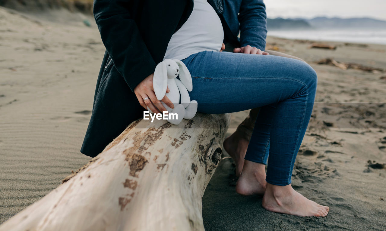 Low section of pregnant woman sitting with man on driftwood at beach